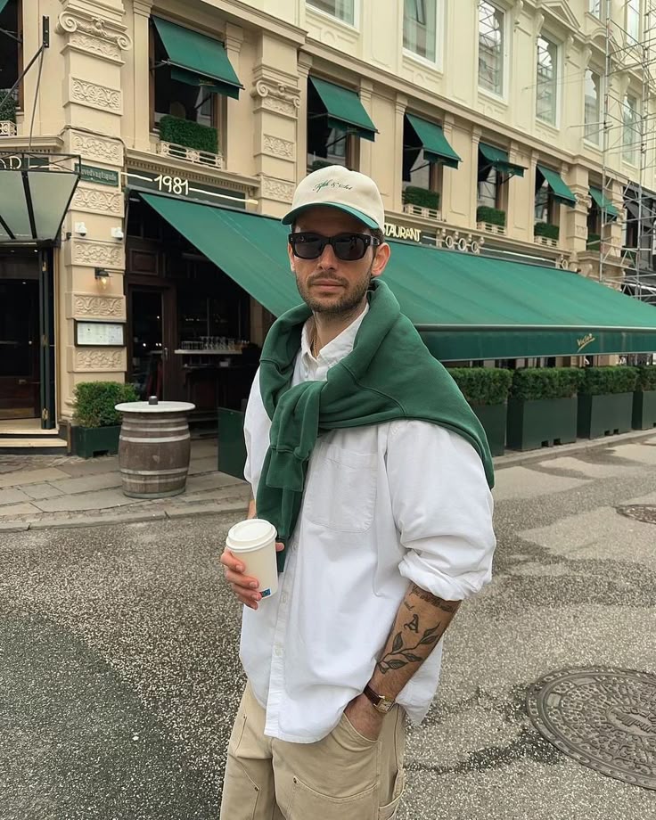 Man in casual outfit holding a coffee cup, standing outside a stylish building with green awnings.