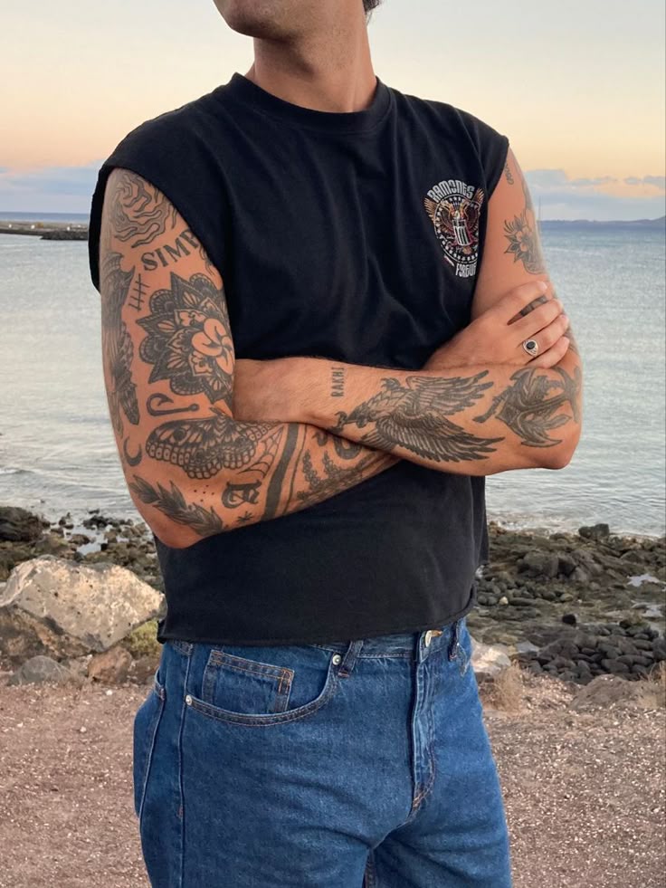 Man with tattooed arms in a black shirt and jeans by the sea during sunset.