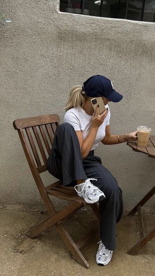Person in casual outfit sitting on outdoor chair, talking on phone, holding iced coffee, wearing cap and sneakers.
