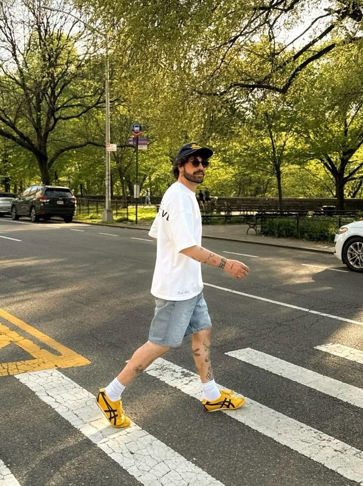 Man in casual summer attire crossing street in park, wearing sunglasses and yellow sneakers.
