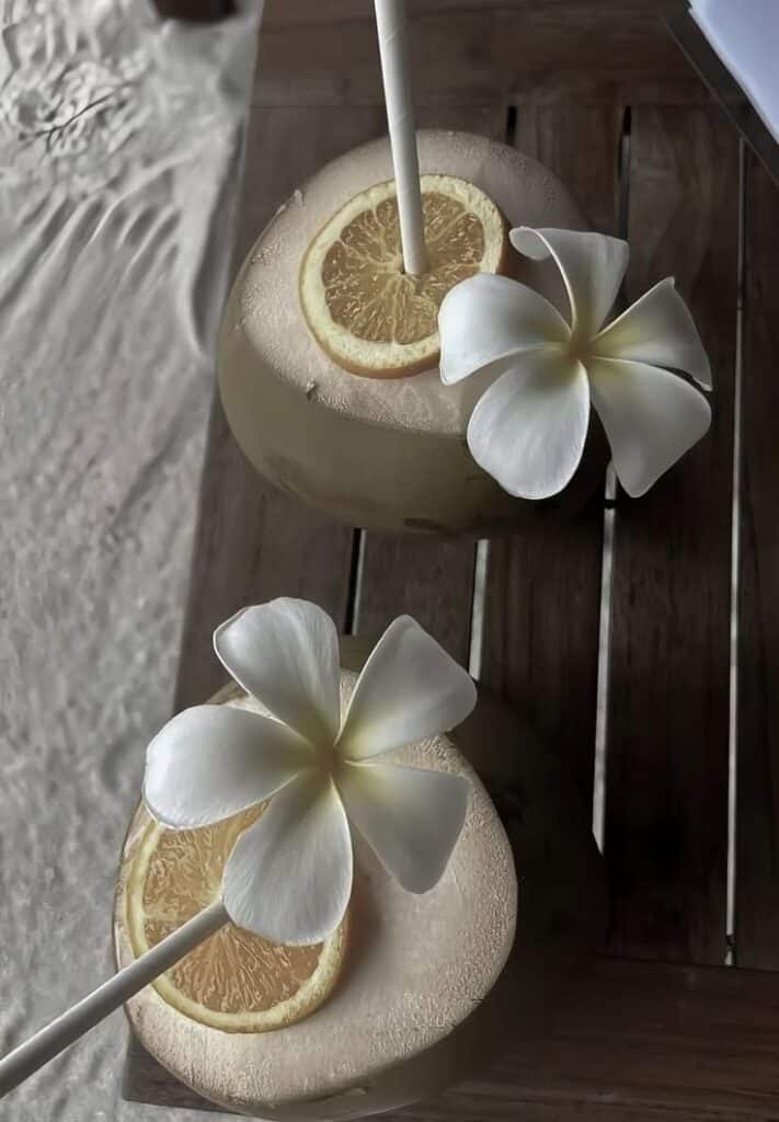 Tropical drinks in coconuts with plumeria flowers and orange slices on a wooden table.