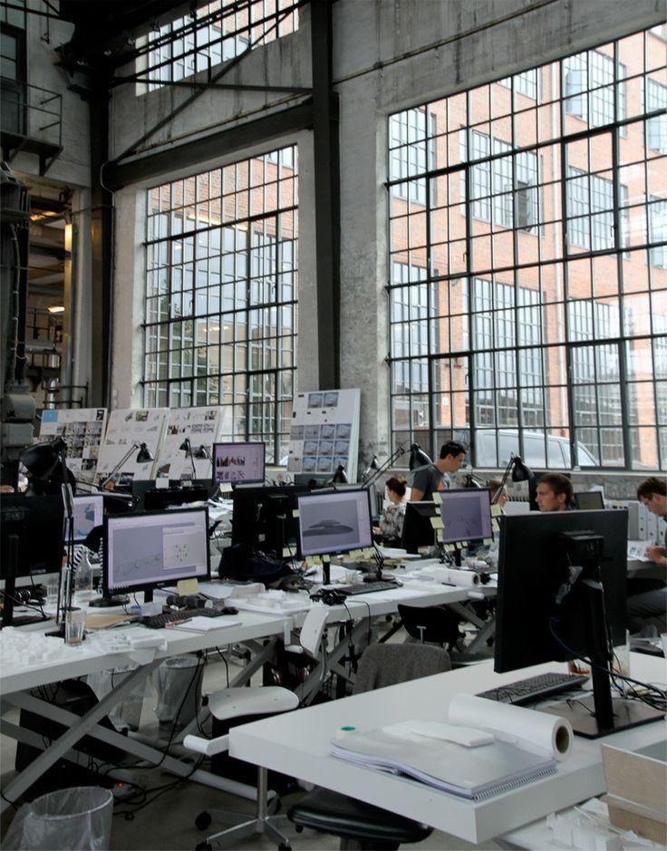 Open office space with large windows, employees working at desks with computers, industrial modern design.