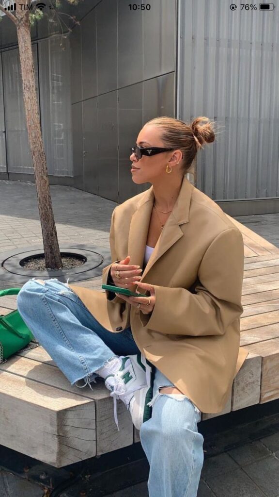 Woman in oversized blazer and jeans sitting on bench outdoors, holding phone with green bag beside her.