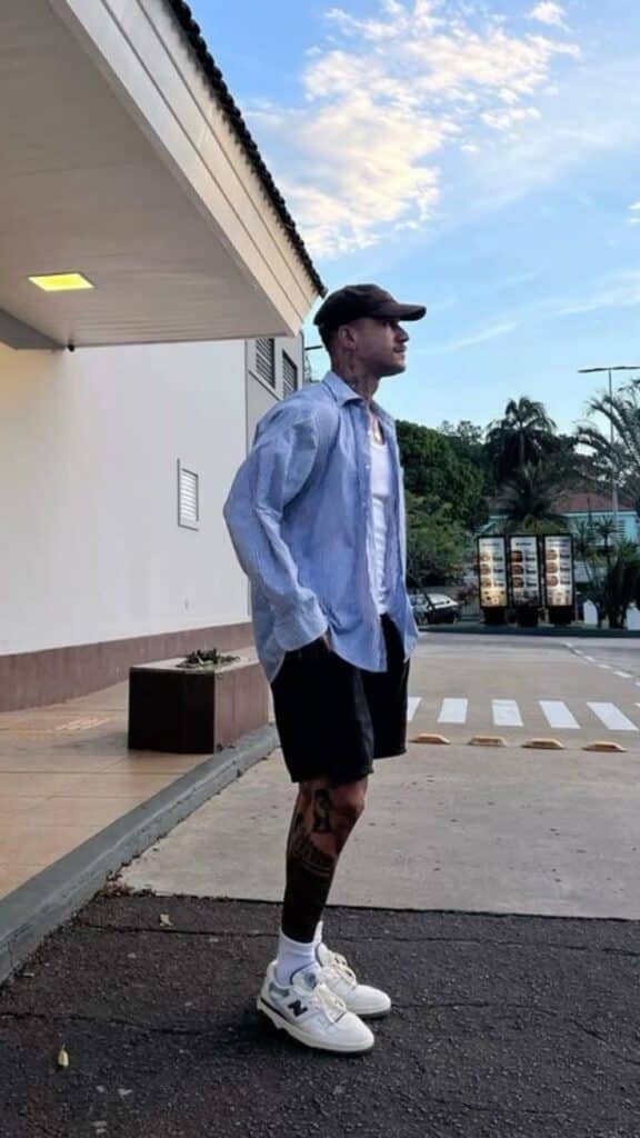 Man in casual outfit standing outside a building with palm trees in the background, wearing sneakers and a cap.
