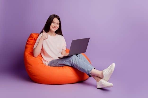 Smiling woman with laptop on orange bean bag gives thumbs-up, against a purple background. Casual home office vibe.