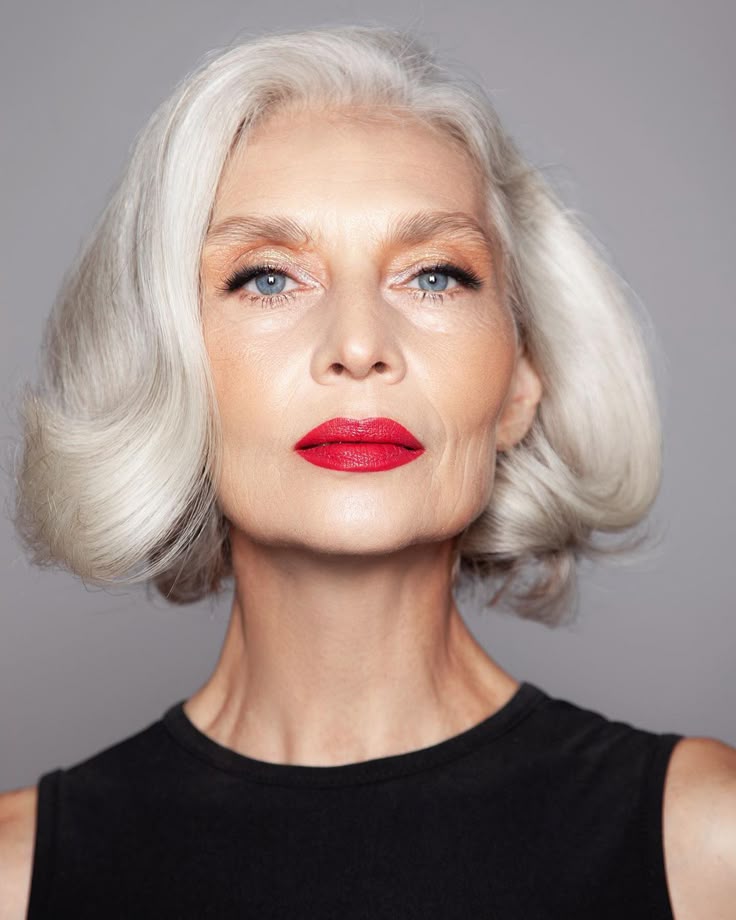 Elegant woman with styled silver hair and red lipstick, wearing a black top on a gray background.
