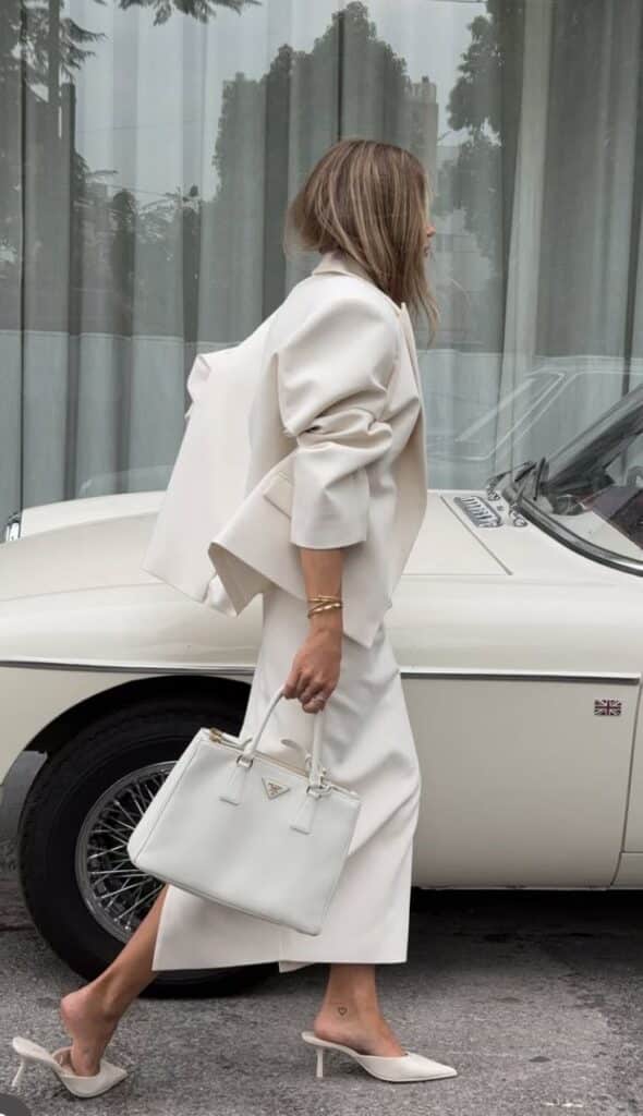 Stylish woman in white outfit and heels with matching handbag walks past vintage car.