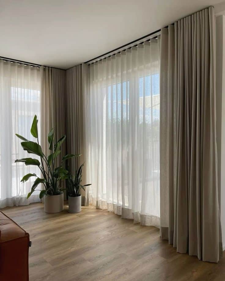 Modern living room with floor-to-ceiling curtains, wooden floor, and potted plants by large windows.