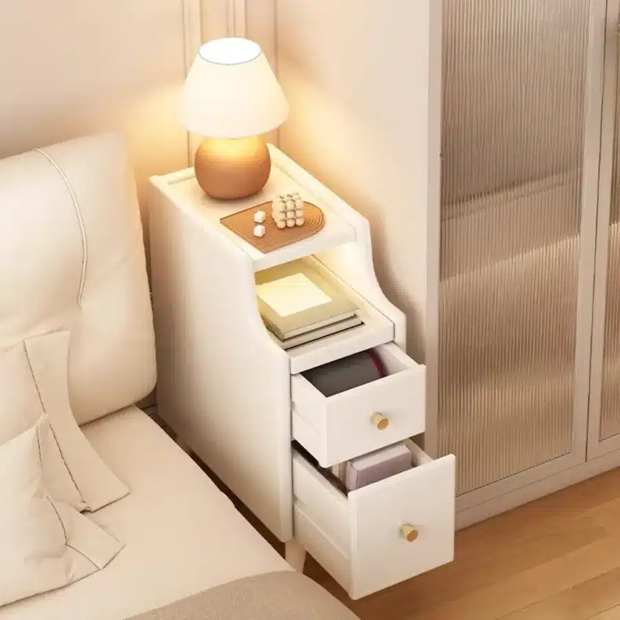Cozy bedroom with a modern white nightstand, lamp, open drawers, and books beside a bed.