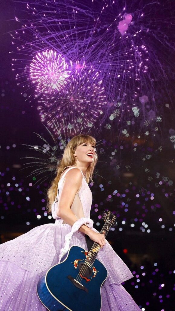 Performer in a purple dress with a blue guitar smiles under vibrant purple fireworks at a nighttime concert.