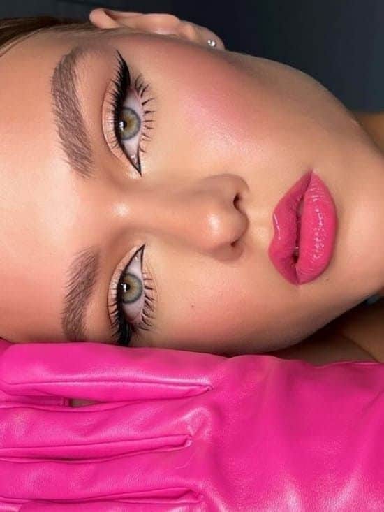 Close-up of a woman with bold makeup and pink lips, resting her face on a pink-gloved hand.