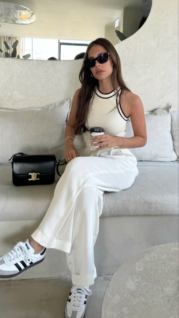 Stylish woman in white outfit with sunglasses holds coffee, sitting on a couch with a black handbag, modern decor.