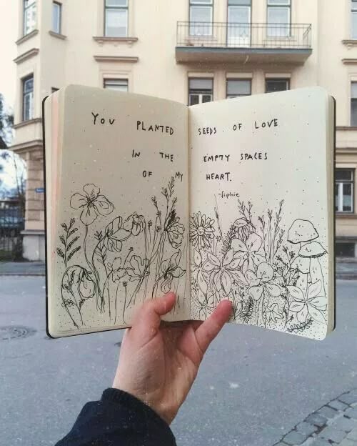 Hand holding an open notebook with floral sketches and a romantic quote about love on a street background.