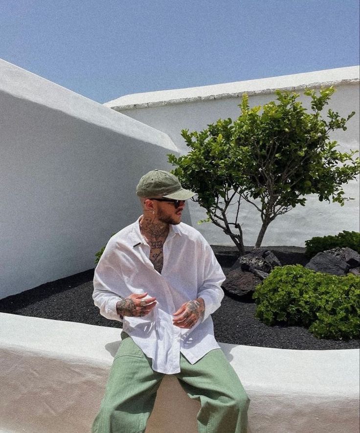 Man in casual attire and cap sits by a tree in a sunny courtyard with white walls. Outdoor fashion and lifestyle.