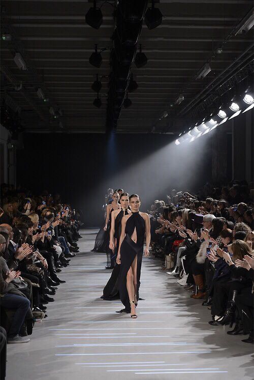 Fashion models in black gowns walk down a well-lit runway, surrounded by an audience at a fashion show.