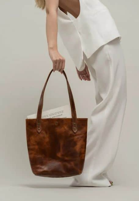 Fashionable woman in white outfit holding a stylish brown leather tote bag with a newspaper inside, minimalist background.