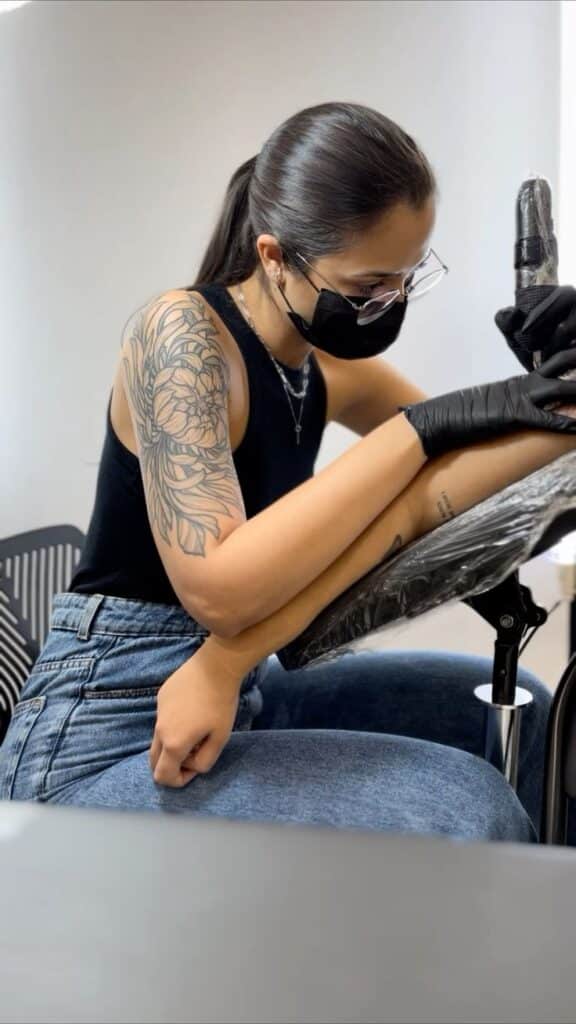 Tattoo artist focused on creating intricate arm design, wearing mask and gloves in studio setting.