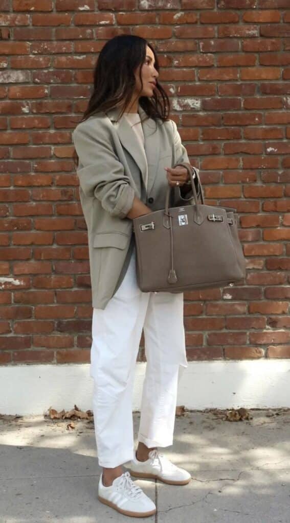 Woman in stylish oversized blazer and white pants holding a gray handbag against a brick wall.