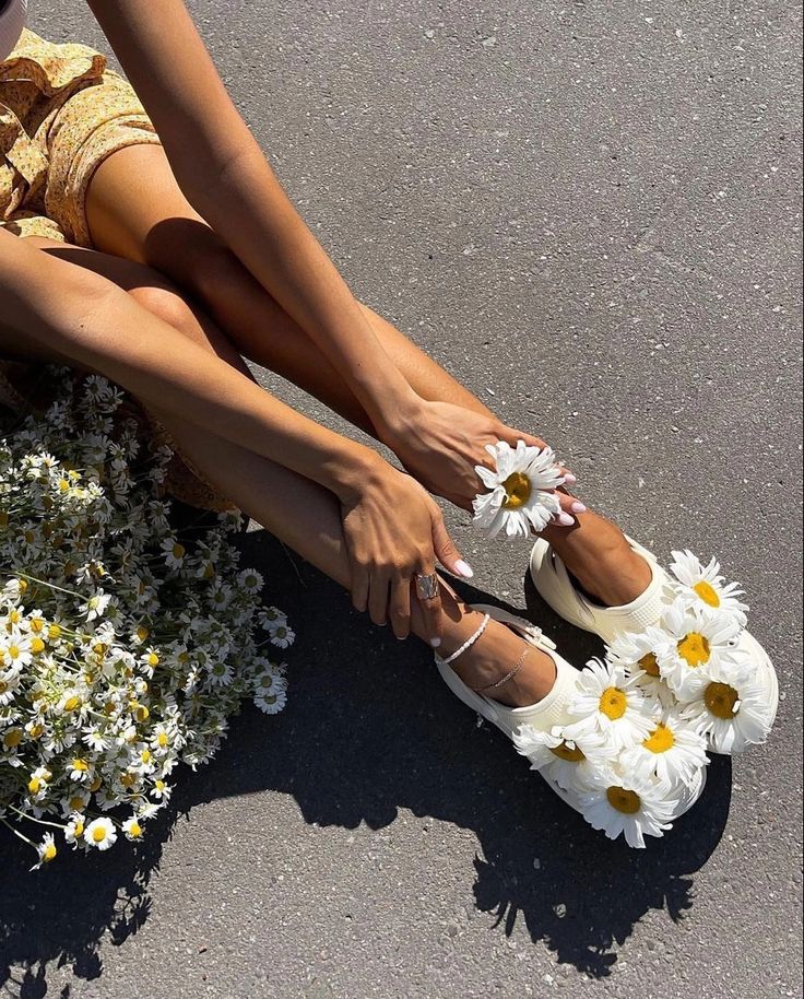 Woman sitting on pavement wearing daisy-decorated shoes, holding a daisy with a bouquet by her side.