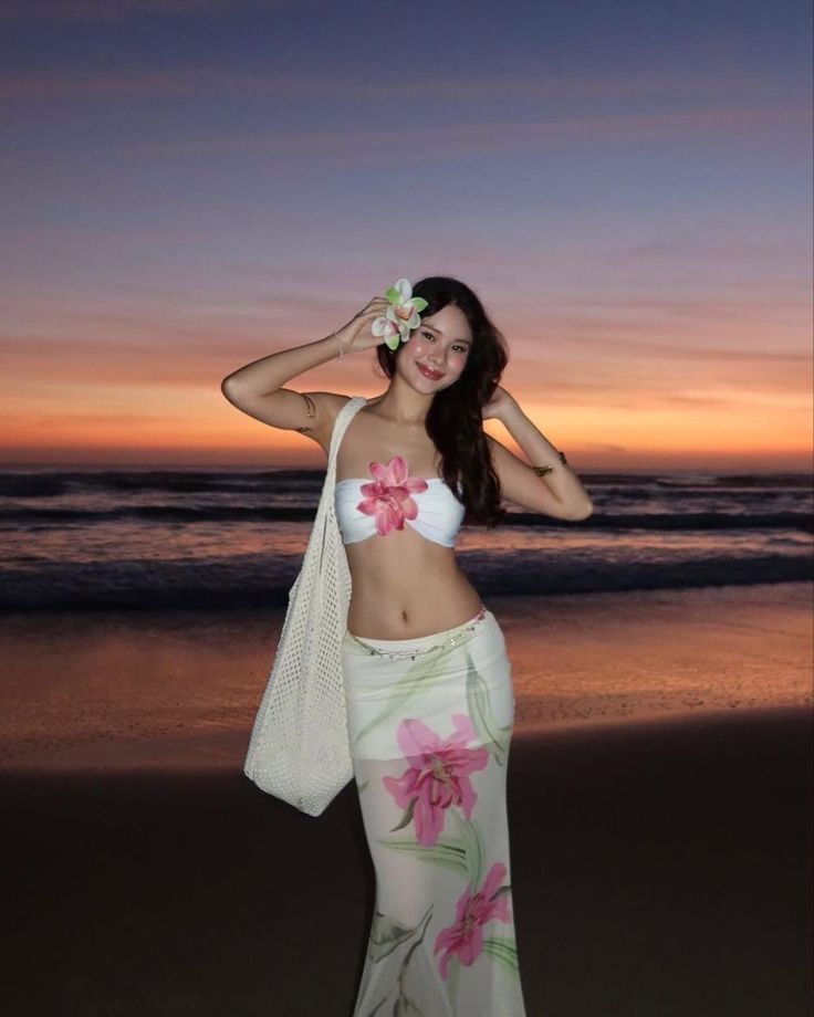 Woman in floral attire posing at sunset beach with a flower in her hair, embodying island vibes and tranquility.
