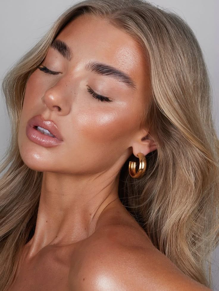 Close-up of a woman with natural makeup, glowing skin, golden hoop earrings, and long blonde hair against a light background.