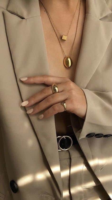 Woman in beige suit with gold jewelry, including necklaces and rings, showcasing elegant style and fashion.