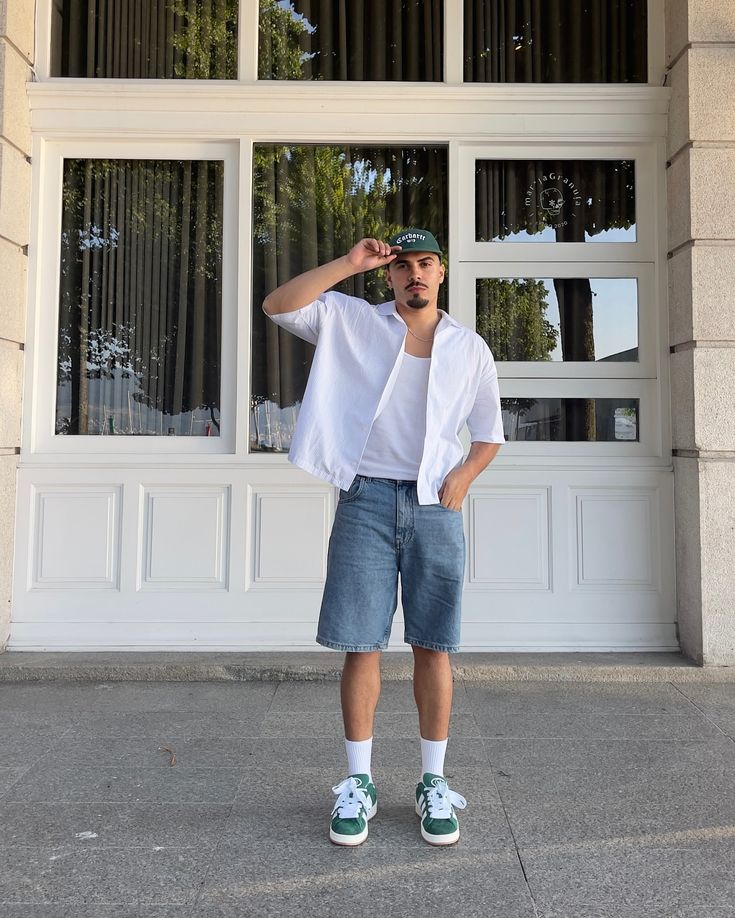 Man in casual outfit with green cap, white shirt, and denim shorts stands in front of windowed building.