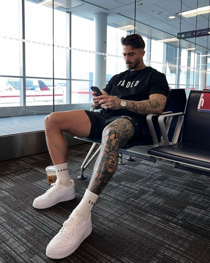 Man with tattoos sitting at airport, wearing a black shirt and white sneakers, using phone.