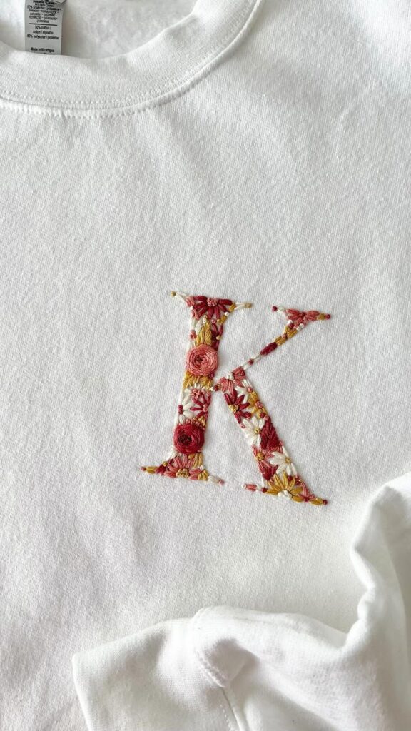 White sweatshirt with floral embroidered letter K design, showcasing intricate red and yellow flower details.