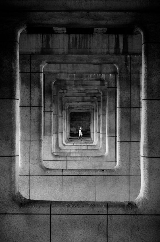 Black and white photo of a person standing in the center of a repetitive architectural tunnel, creating a symmetrical effect.