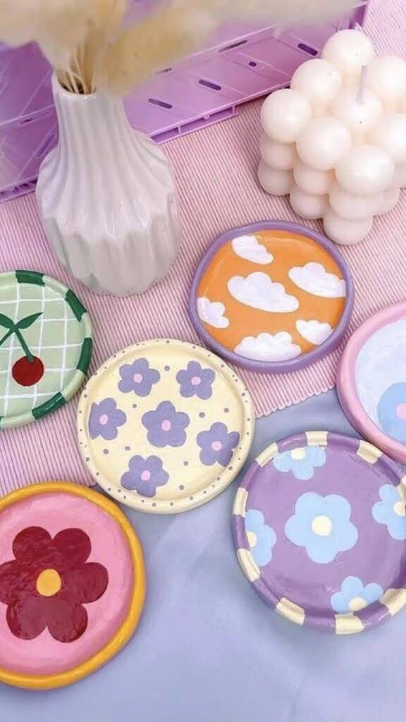 Colorful handmade coasters with floral and cloud patterns on a pink surface, alongside a vase and bubble candle.
