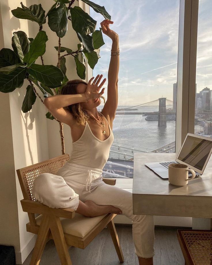 Woman doing yoga stretches at home office with city bridge view through window.