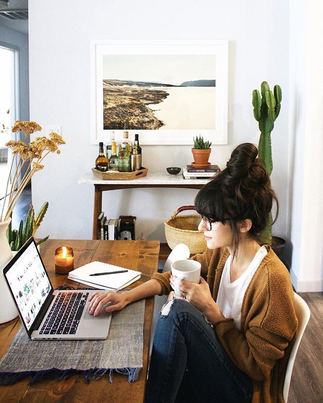 Cozy workspace with a woman holding a mug, using a laptop, surrounded by decor and plants.