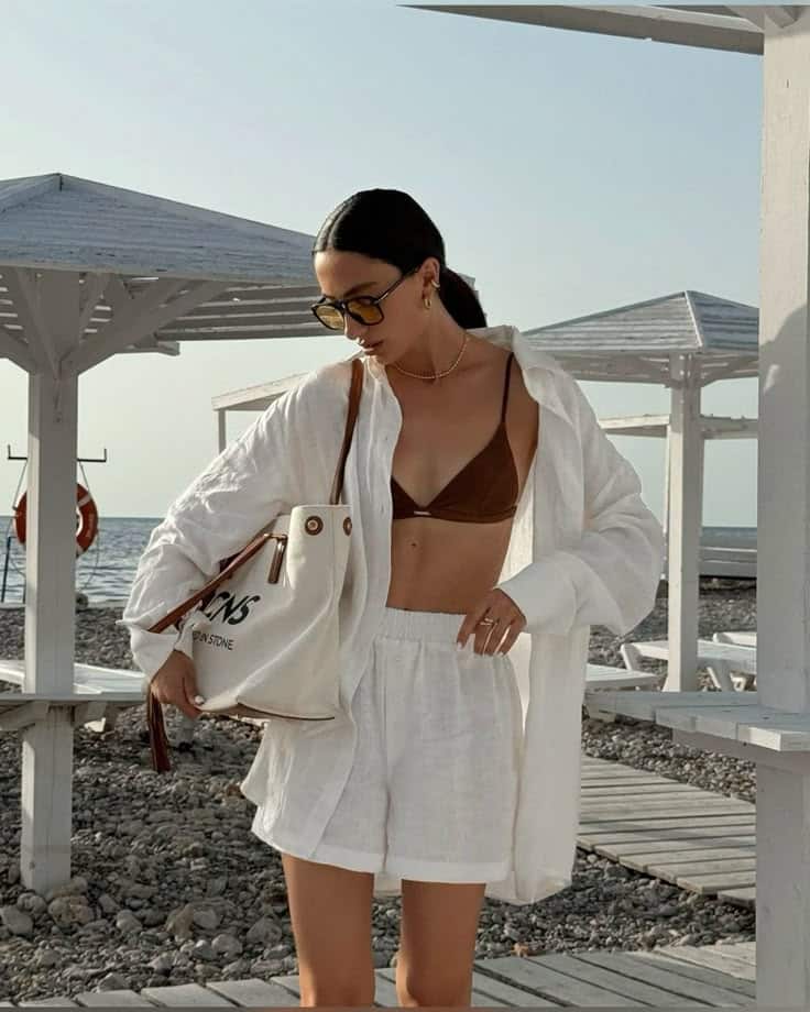 Woman in white beach attire, sunglasses, and a tote bag walking on a pebble beach with wooden huts.