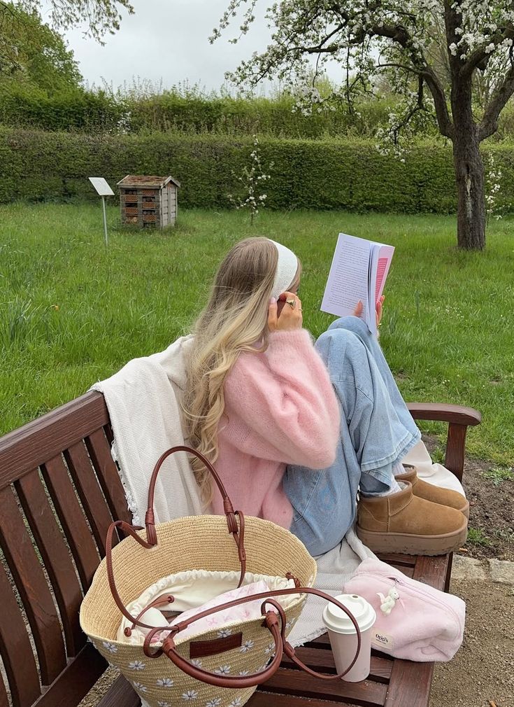 1f4cd860-613b-49ed-86f1-3c1f7df82c23 Person in pink sweater reading on a bench in a garden, with a straw bag and coffee nearby. | Sky Rye Design Person in pink sweater reading on a bench in a garden, with a straw bag and coffee nearby.