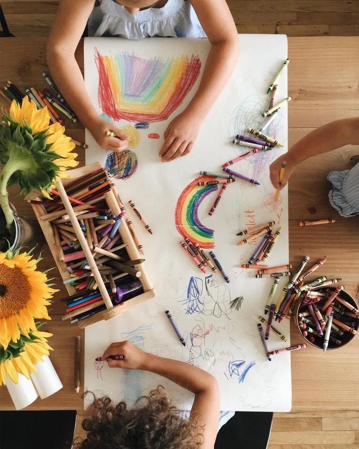 1f81045b-de66-4740-ab21-d4f9d6dbb1a6 Children drawing rainbows and shapes with crayons on a table, surrounded by sunflowers and art supplies. | Sky Rye Design Children drawing rainbows and shapes with crayons on a table, surrounded by sunflowers and art supplies.