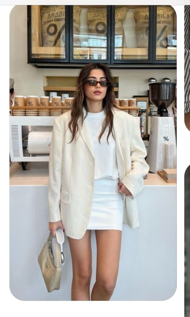 Fashionable woman in white blazer and mini skirt with sunglasses, standing in a modern coffee shop.