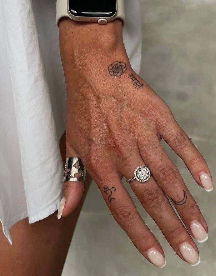 Hand with tattoos, wearing a silver ring and a diamond ring, and a smartwatch on the wrist against a neutral background.
