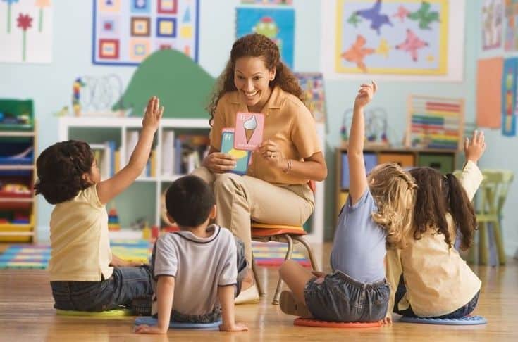 Teacher engaging with young children in a colorful classroom, holding flashcards during an interactive learning session.