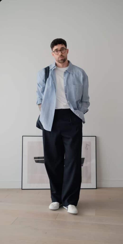Man in casual outfit with blue shirt, white tee, and black pants standing indoors with framed art in background.