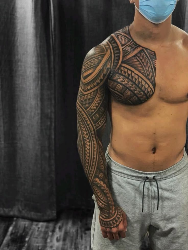 Man with intricate Polynesian tattoo sleeve on arm and chest, wearing a mask, standing in a studio setting.
