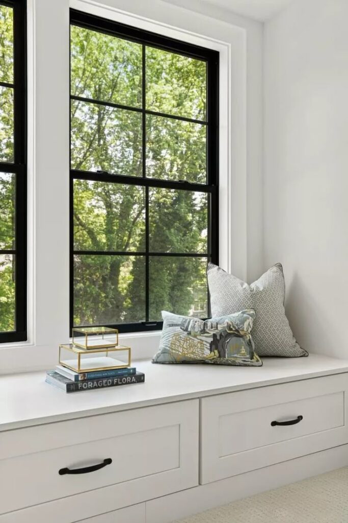Cozy window seat with decorative pillows and books, overlooking lush greenery through black-framed windows.