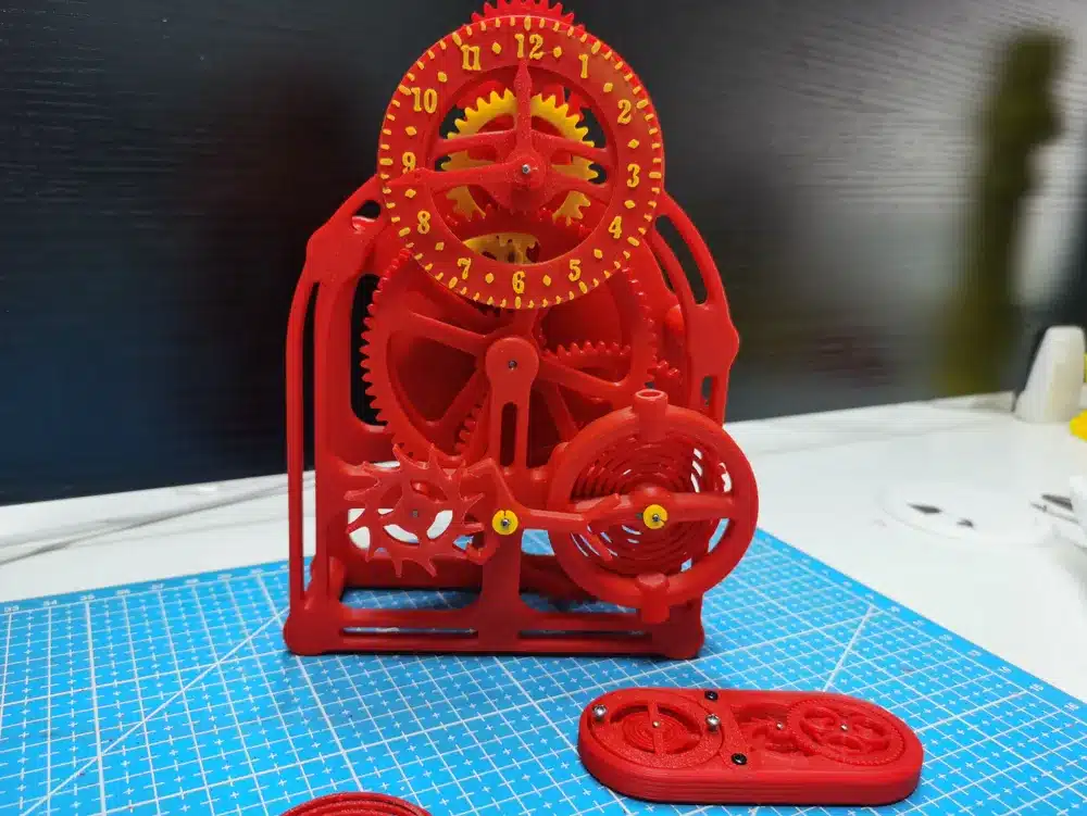 3D-printed red gear mechanism with clock design on a blue cutting mat, showcasing intricate machinery.