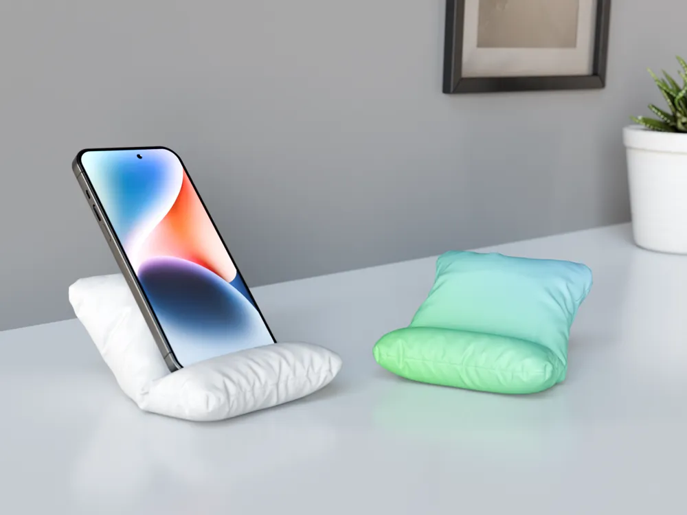 Smartphone on a mini pillow stand on desk, next to another empty pillow holder, near plant and framed picture.