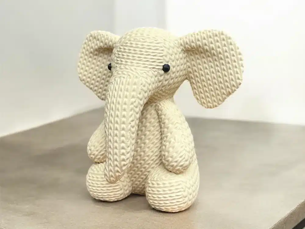 Knit elephant toy sitting on a shelf, made of cream-colored yarn, with large ears and black button eyes.