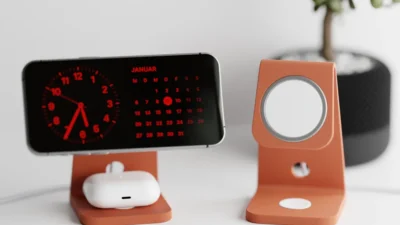 Smartphone on a stand with digital clock and calendar display, AirPods below; modern desk setup with plant in background.