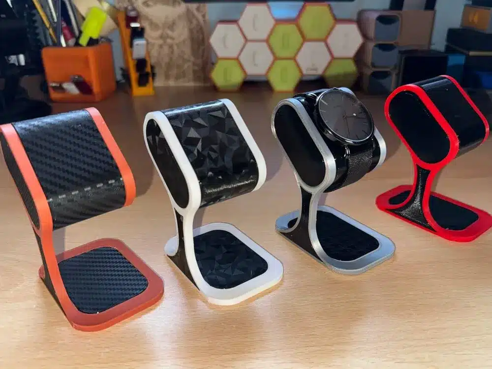 Four stylish 3D-printed watch stands in various colors, with one holding a black watch on a wooden desk.
