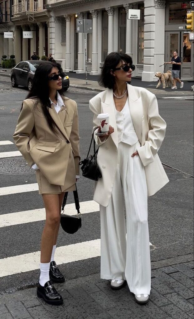 Two fashionable women in stylish blazers and sunglasses crossing a city street.