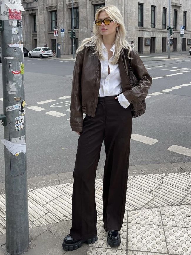 Stylish woman in urban setting wearing a brown leather jacket, white shirt, and sunglasses at a city street corner.