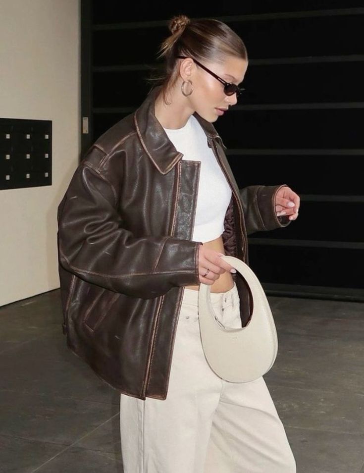 Fashionable woman in brown leather jacket, sunglasses, and white pants holding a trendy bag.